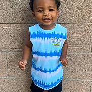 Christofer is registered to the contest to win money with this photo: toddler, child, smiling, standing, sleeveless_shirt, tie_dye, black_shorts, sneakers, urban, brick_wall, concrete, happy, portrait, casual_clothing, outdoor, person, cute, short_hair, playful, feet