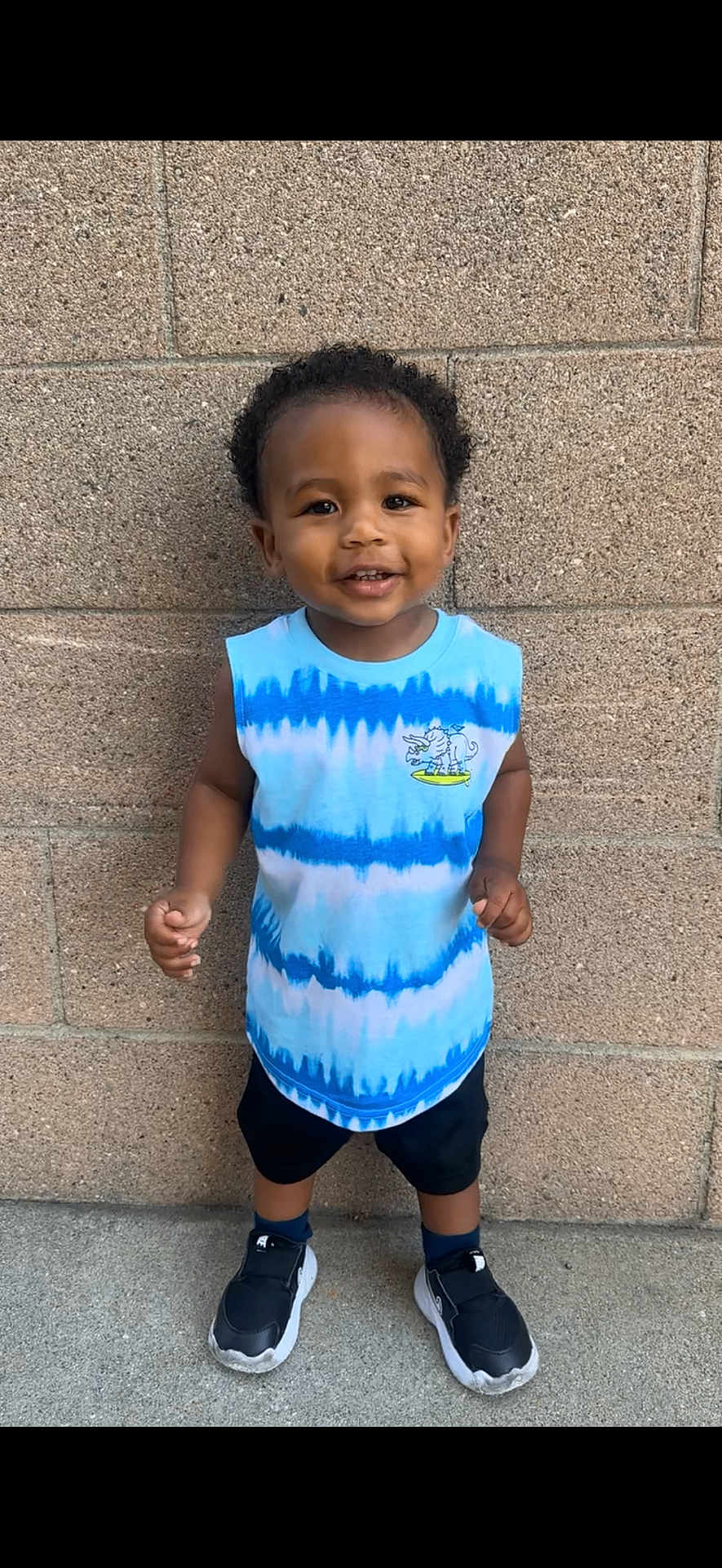 Christofer is registered to the contest to win money with this photo: toddler, child, smiling, standing, sleeveless_shirt, tie_dye, black_shorts, sneakers, urban, brick_wall, concrete, happy, portrait, casual_clothing, outdoor, person, cute, short_hair, playful, feet