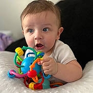 Levi is registered to the contest to win money with this photo: baby, child, toy, colorful, plastic, blue_eyes, curious, cushion, blanket, indoor, face, hand, expression, infant, soft, playing, person, closeup, cute, young