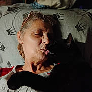 Midnight joined the competition — help win amazing prizes! woman, older_woman, cat, black_cat, pet, licking, kissing, affectionate, cuddle, pillow, paw_print, glasses_on_head, indoor, closeup, portrait, relaxed, night_time, paw, collar, sofa