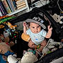 baby, child, playpen, toys, stuffed_animals, hat, cap, drool, indoor, bookshelf, books, colorful, cute, curious, sitting, person, infant, clothing, baby_toys, soft_toys