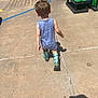 toddler, child, back_view, blue_romper, rain_boots, shark_pattern, sidewalk, concrete, sunny_day, outdoor, cleaning_supplies, plastic_bottles, parking_lot, shadow, walking, casual_clothing, short_hair, daylight, young_child, person