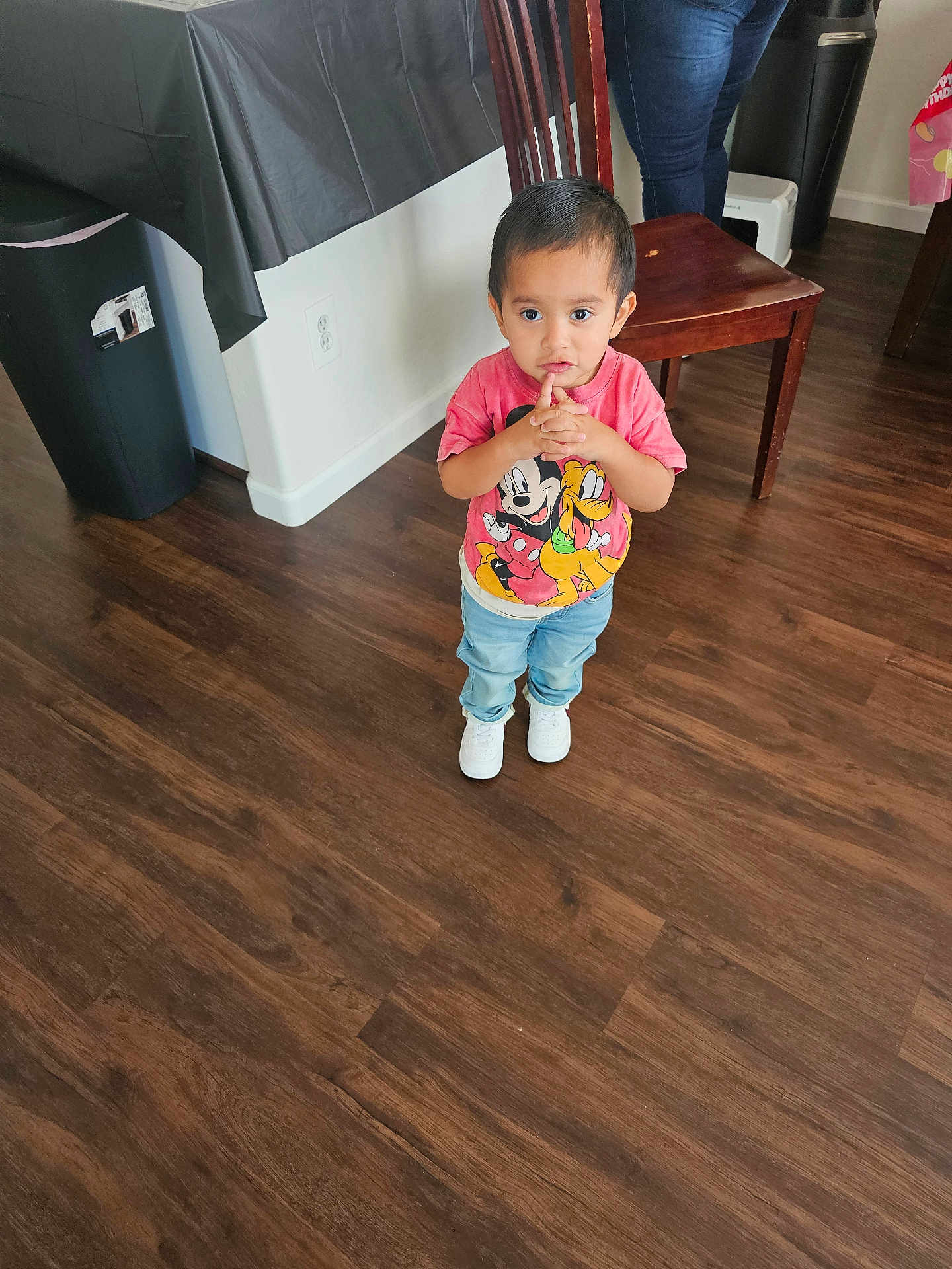 Ezekiel joined the competition — help win amazing prizes! toddler, child, indoor, floor, wooden_floor, jeans, tshirt, mickey_mouse, pluto, white_shoes, chair, table, black_tablecloth, person, standing, looking_up, hands_together, expression, adult, home