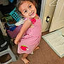 child, girl, smiling, red_dress, checkered_pattern, barefoot, indoor, carpet, clutter, shoe, hanger, lamp, closet, toy, person, cute, happy, posing, room, playful