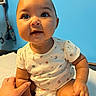 Paloma is registered to the contest to win money with this photo: baby, child, infant, cute, smiling, sitting, onesie, diaper, hand, arm, blue_background, curious, chubby_cheeks, indoor, portrait, skin, person, young_child, happy, bright