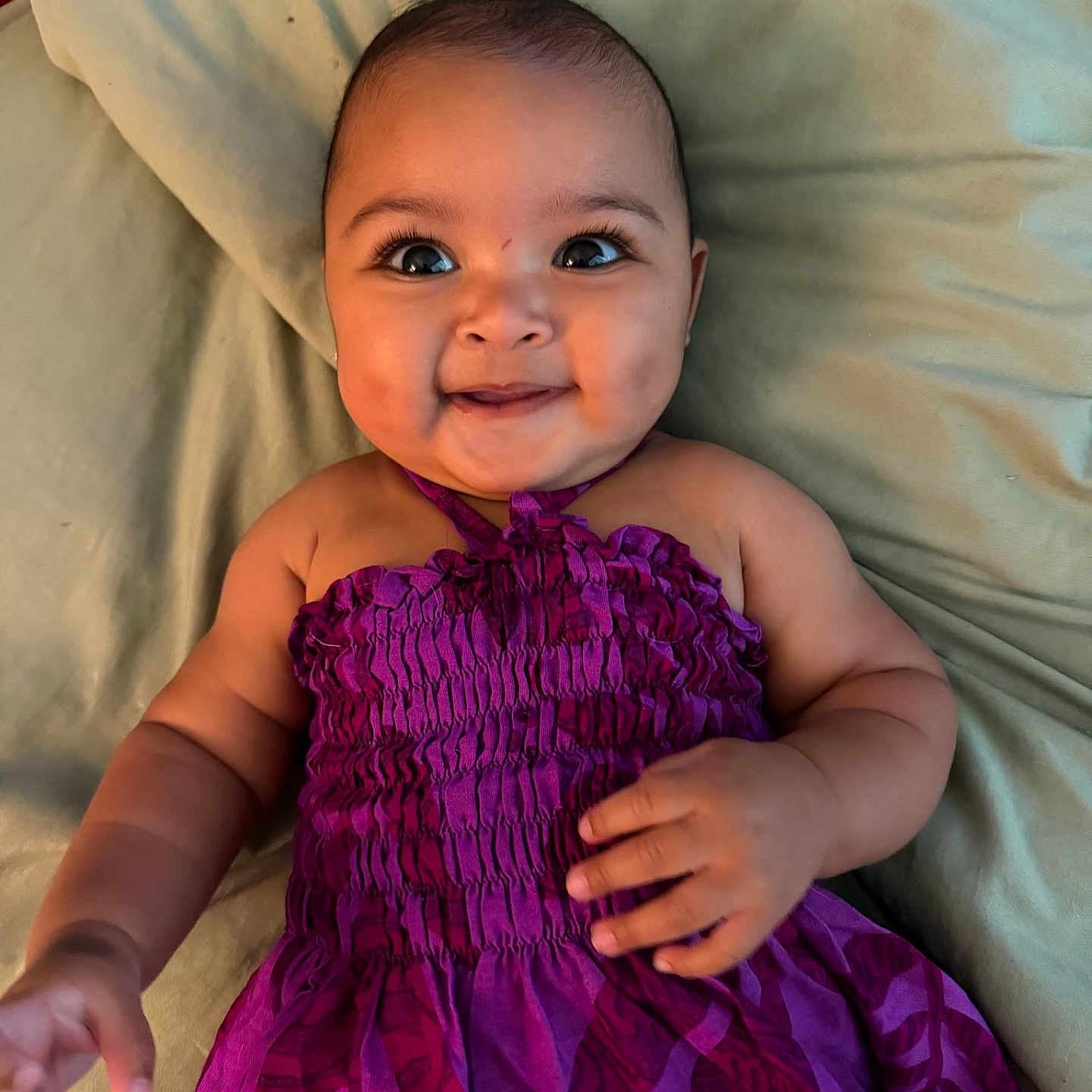 Paloma is registered to the contest to win money with this photo: baby, child, smiling, purple_dress, lying_down, blanket, cute, happy, portrait, infant, person, face, skin, hand, sleepwear, indoors, closeup, young, cheerful, bright