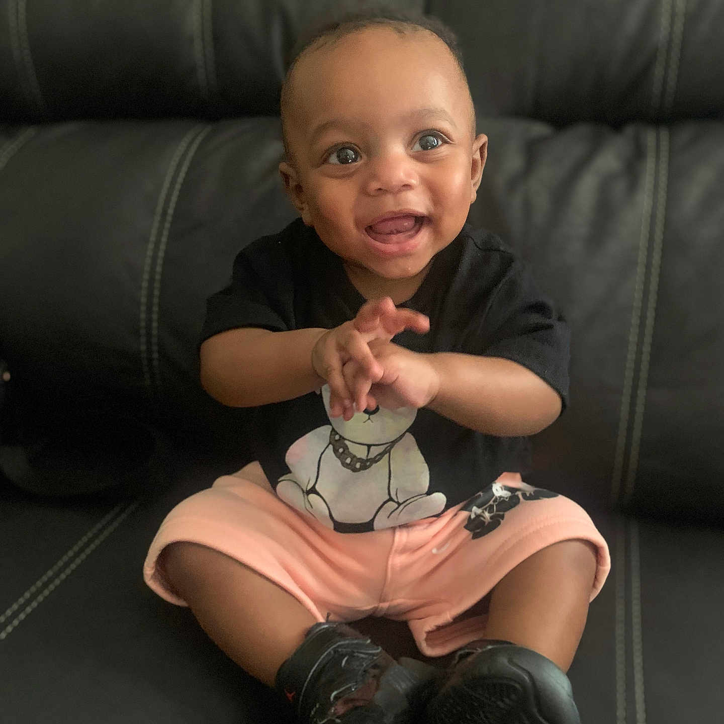 Jay R is registered to the contest to win money with this photo: baby, child, smiling, sitting, black_tshirt, teddy_bear, shorts, black_shoes, leather_couch, happy, clapping, indoor, cute, portrait, person, infant, young_child, expression, casual_clothing, fun