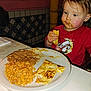 toddler, child, messy_face, food, rice, beans, quesadilla, plate, red_shirt, cartoon_character, restaurant, table, eating, meal, indoor, high_chair, window, tile_wall, yellow_pole, napkin