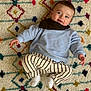baby, child, smiling, blue_sweater, striped_pants, white_socks, patterned_rug, colorful, indoor, cute, happy, portrait, infant, person, laying_down, cozy, small_hands, small_feet, head, face