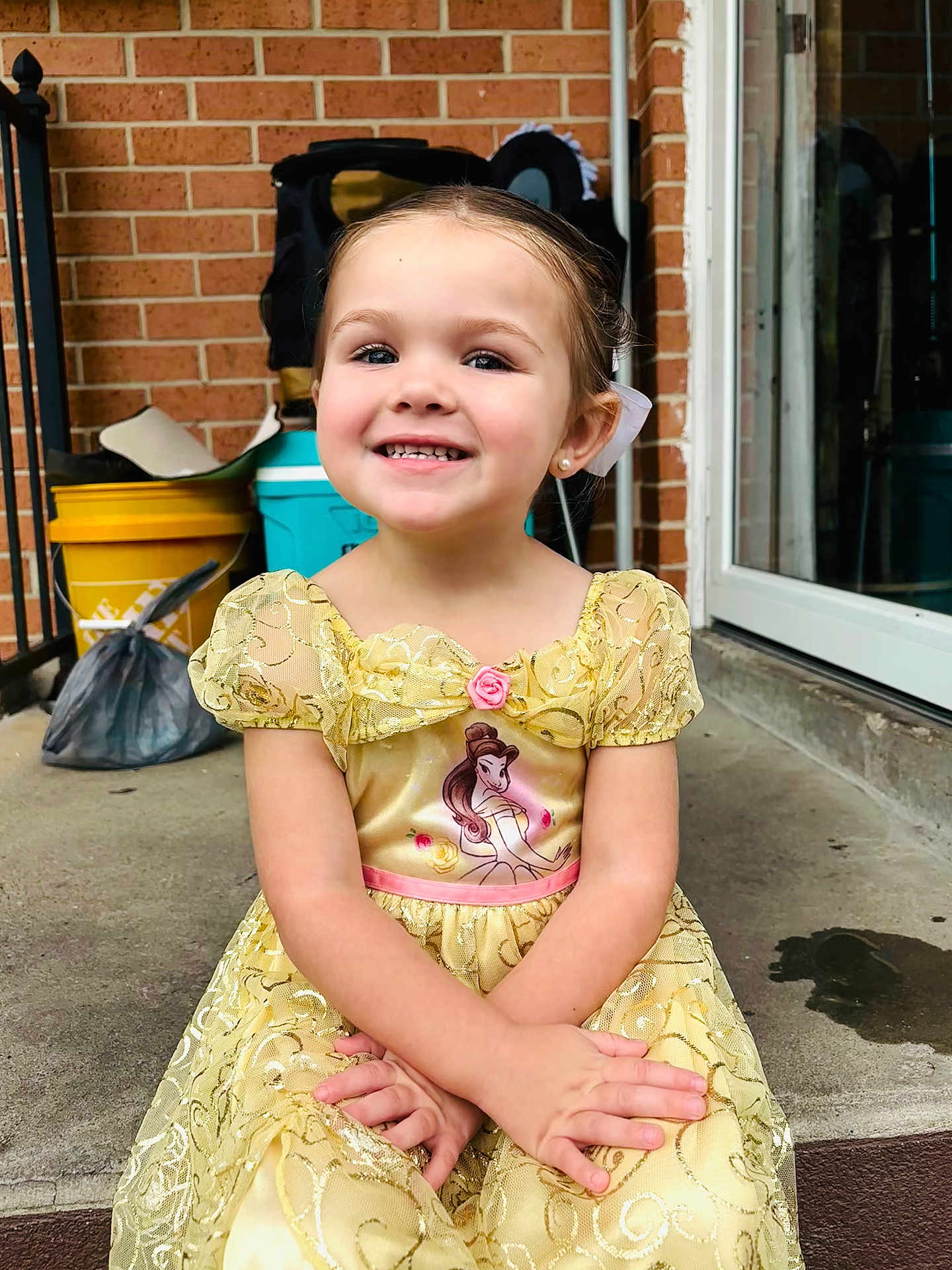 Oakley is registered to the contest to win money with this photo: child, girl, smile, dress, yellow_dress, princess, costume, sitting, concrete, brick_wall, outdoor, happy, portrait, crossed_arms, young, person, casual, daylight, background_objects, earrings