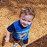 toddler, child, smiling, curly_hair, blue_shirt, paw_patrol, shoes, wood_chips, outdoor, sunlight, shadow, playground, happy, person, casual_clothing, standing, cute, daylight, skin, young_child