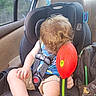 child, car_seat, toy, curly_hair, blue_shirt, shorts, sneakers, car_interior, window, seatbelt, bag, elephant_pattern, hand, leg, head, side_view, greenery, black_car_seat, red_toy, seat