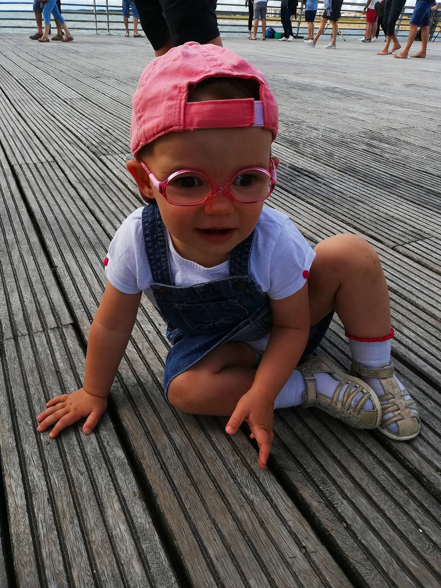 Maëlys participe au concours pour gagner de l'argent avec cette photo : boardwalk, child, deck, floor, flooring, headgear, headwear, person, sitting, smile, sunglasses, toddler, vacation, walkway, wood