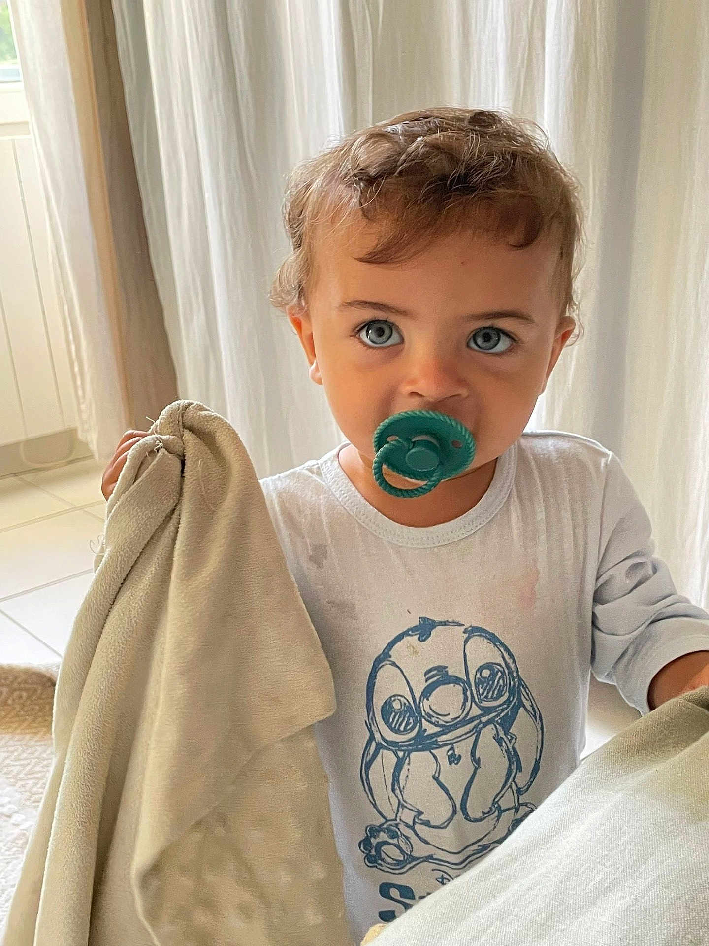 Nesta participe au concours pour gagner de l'argent avec cette photo : toddler, child, pacifier, blanket, indoor, curly_hair, blue_eyes, white_shirt, cartoon_print, soft_light, curtain, floor_tiles, blanket_clutching, closeup, portrait, cute, baby, young_child, comfort, casual_clothing