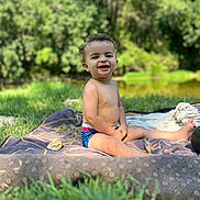 Nesta participe au concours pour gagner de l'argent avec cette photo : child, toddler, smiling, outdoor, grass, blanket, curly_hair, sunlight, nature, picnic, happy, bare_chest, shorts, greenery, park, sitting, face, daylight, relaxation, playful