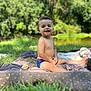 child, toddler, smiling, outdoor, grass, blanket, curly_hair, sunlight, nature, picnic, happy, bare_chest, shorts, greenery, park, sitting, face, daylight, relaxation, playful