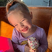 Scarlett is registered to the contest to win money with this photo: child, girl, ice_cream, sprinkles, cone, smile, happy, indoor, wooden_bench, purple_shirt, food, dessert, sunlight, face, hands, table, pacifier, hair_tie, cute, messy_eating
