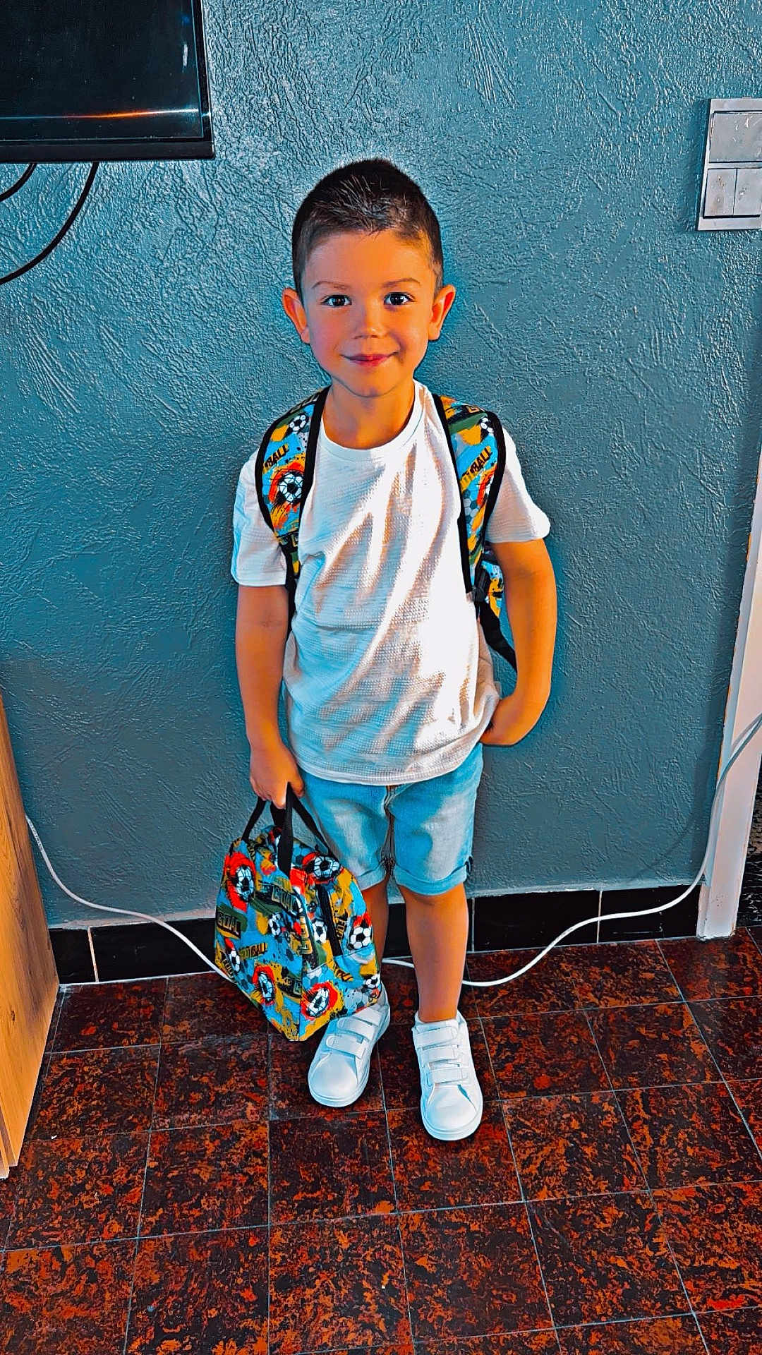 Manelyo participe au concours pour gagner de l'argent avec cette photo : child, boy, backpack, lunch_bag, white_tshirt, shorts, sneakers, indoor, floor, wall, smiling, standing, person, casual_clothing, school_bag, happy, portrait, young, ready, colorful
