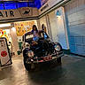 child, car, vintage_car, gas_station, gas_pump, sign, license_plate, indoor, lighting, pose, smile, vehicle, black_car, floor, door, wall, person, automobile, headlamp, retro