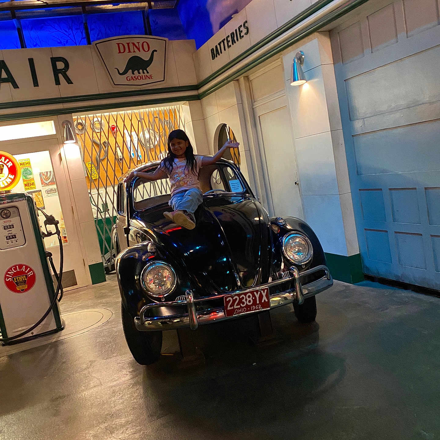 Renata Portales is registered to the contest to win money with this photo: child, car, vintage_car, gas_station, gas_pump, sign, license_plate, indoor, lighting, pose, smile, vehicle, black_car, floor, door, wall, person, automobile, headlamp, retro