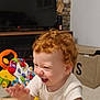 baby, carpet, child, clothing, curly_hair, furniture, hand, happy, indoor, laughing, living_room, person, playful, red_hair, smiling, storage_box, table, television, toddler, toy