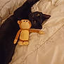 black_cat, plush_toy, monkey_toy, bed, blanket, stretching, pet, indoor, cute, animal, feline, cozy, soft_toy, resting, relaxed, companion, playful, yellow_eyes, fur, sleepy