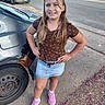brown_hair, brown_shirt, car, child, daylight, denim_shorts, fashion, flowers, girl, hands_on_hips, long_hair, outdoor, pink_shoes, portrait, residential_area, sidewalk, smiling, standing, street, vehicle
