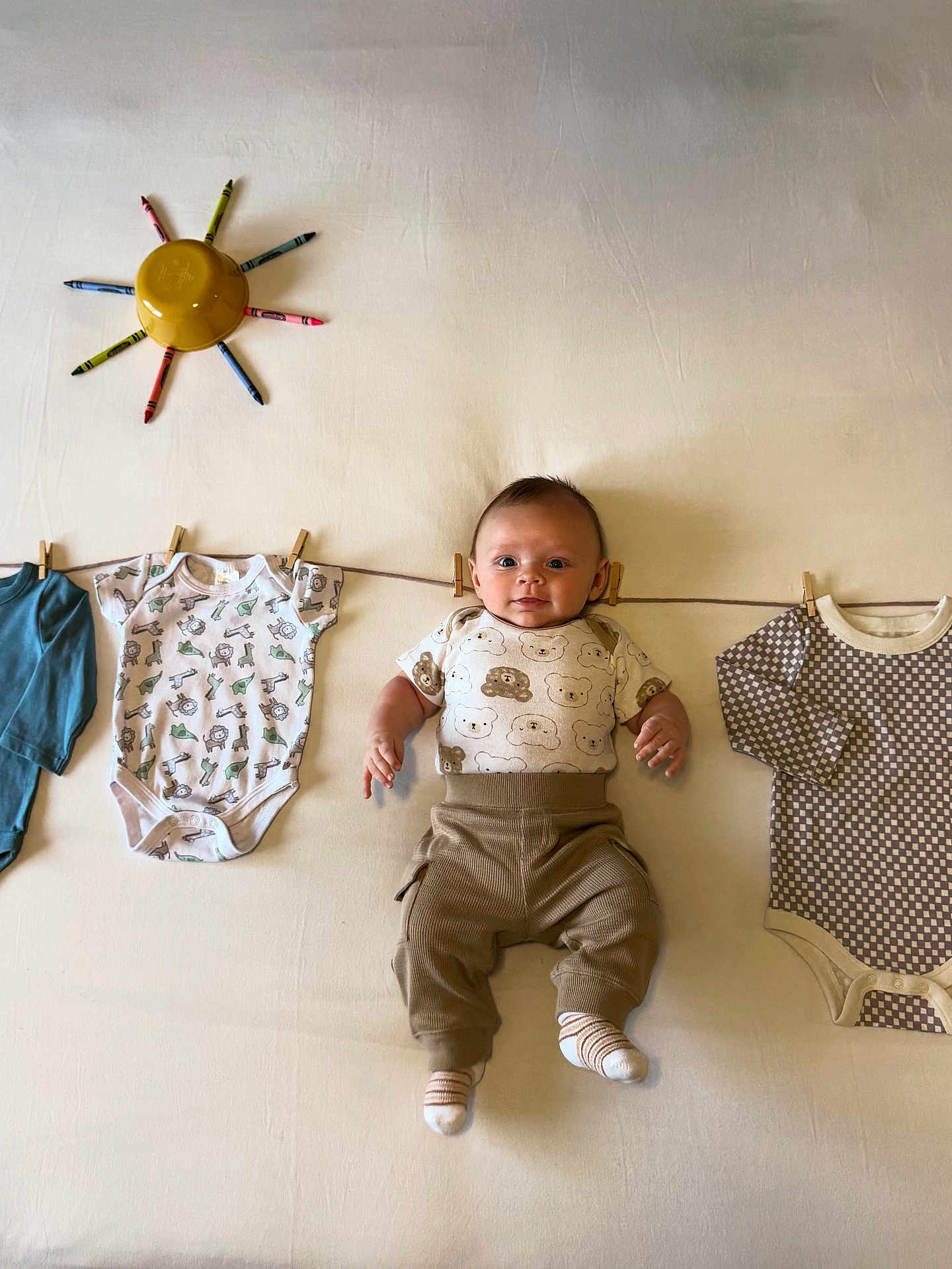 Rowan is registered to the contest to win money with this photo: baby, infant, clothing, onesie, pants, socks, crayons, bowl, clothesline, clothespins, face, smile, cute, indoor, flat_lay, creative, playful, child, young, soft_lighting