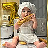 baby, child, chef_hat, apron, bread, baguette, kitchen, countertop, gold_balloon, number_7, rice_cooker, microwave, tile_wall, barefoot, sitting, food, cute, portrait, indoor, playful