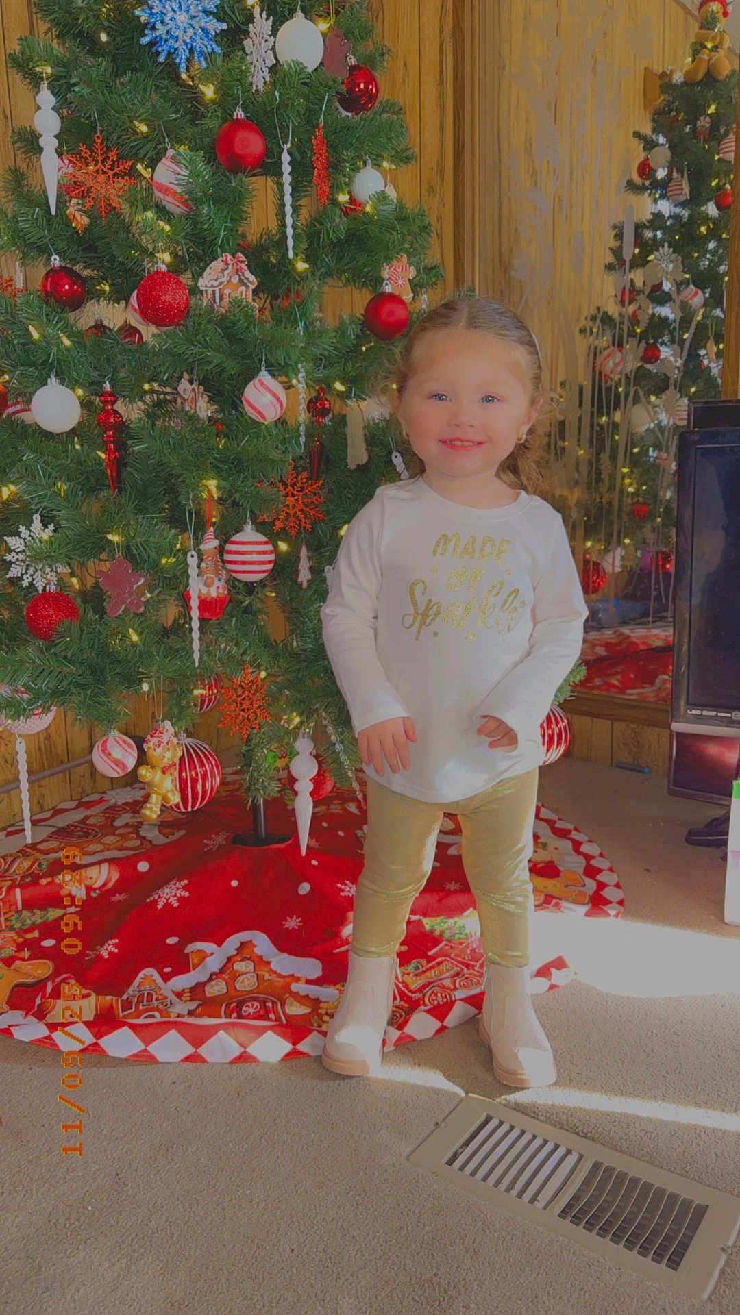 Bellarose is registered to the contest to win money with this photo: child, toddler, christmas_tree, holiday_decorations, ornaments, red_balls, white_balls, snowflakes, gold_leggings, white_shirt, boots, carpet, vent, smiling, indoor, festive, lights, wood_paneling, mirror, television