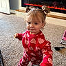 toddler, child, indoor, carpet, red_clothing, hearts_pattern, hair_buns, snack, fireplace, home, floor, cute, person, young_child, smiling, playing, cozy, warm, living_room, curly_hair
