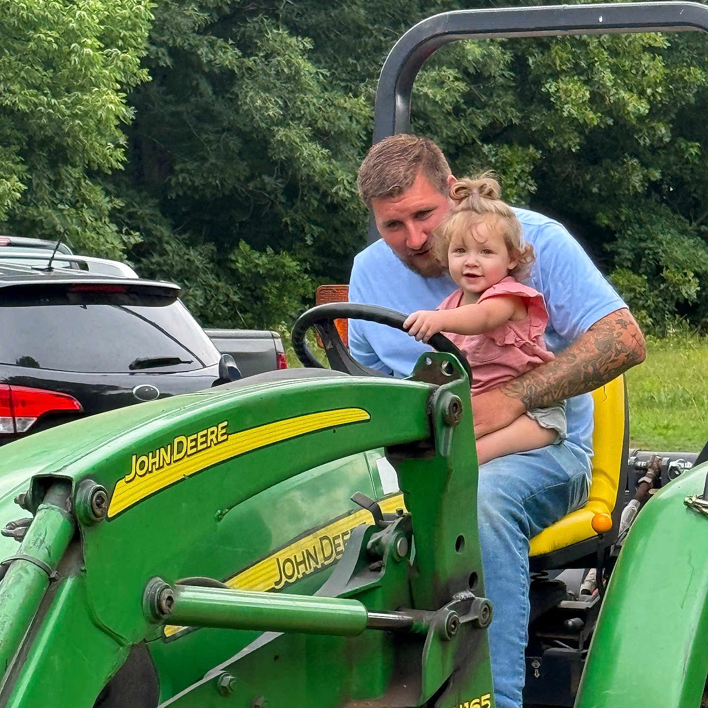 Kaylie joined the competition — help win amazing prizes! child, toddler, man, tractor, john_deere, green, outdoor, nature, trees, vehicle, smile, tattoos, casual_clothing, steering_wheel, yellow_seat, parked_cars, grass, daylight, happy, family