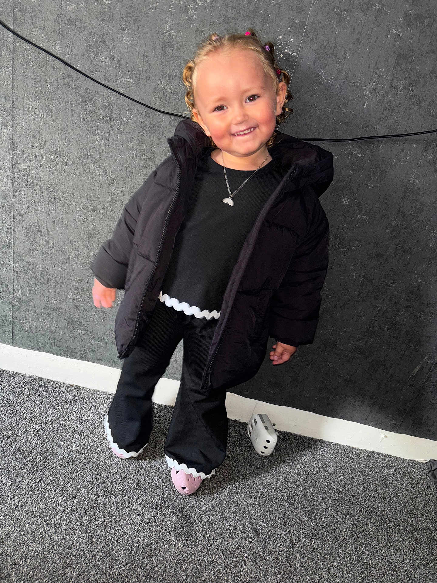 Paisley joined the competition — help win amazing prizes! toddler, child, smiling, curly_hair, black_jacket, black_pants, pink_crocs, carpet, wall, indoor, cute, happy, standing, necklace, hair_clips, fashion, portrait, person, clothing, floor