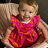 baby, child, smiling, pink_dress, orange_pattern, cushioned_chair, indoor, portrait, happy, cute, person, sitting, infant, playful, face, blue_eyes, short_hair, soft_fabric, home, bright_colors