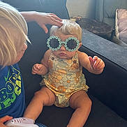 Brynleigh is registered to the contest to win money with this photo: child, toddler, baby, sunglasses, flower_shaped_glasses, romper, couch, indoor, window, hand, head_touch, footwear, shoe, blond_hair, interaction, casual_clothing, seated, furniture, cute, playful