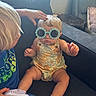 child, toddler, baby, sunglasses, flower_shaped_glasses, romper, couch, indoor, window, hand, head_touch, footwear, shoe, blond_hair, interaction, casual_clothing, seated, furniture, cute, playful
