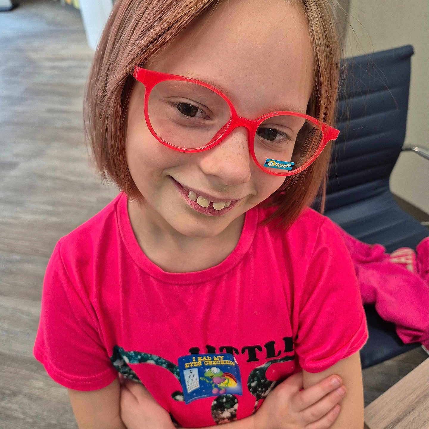 Gabriella is registered to the contest to win money with this photo: child, smile, glasses, red_glasses, pink_shirt, short_hair, indoor, chair, crossed_arms, happy, person, face, freckles, wood_floor, table, casual_clothing, seated, modern_interior, young, cute
