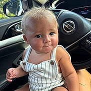 Kohen is registered to the contest to win money with this photo: baby, child, infant, person, car_interior, steering_wheel, nissan_logo, window, greenery, romper, striped_clothing, skin, hair, face, eyes, hand, lap, seat, casual, portrait