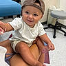 Kohen is registered to the contest to win money with this photo: baby, child, cap, white_shirt, smiling, indoor, hospital, medical_chair, stool, floor, person, hand, legs, cute, infant, holding_hand, casual_clothing, skin, face, seated