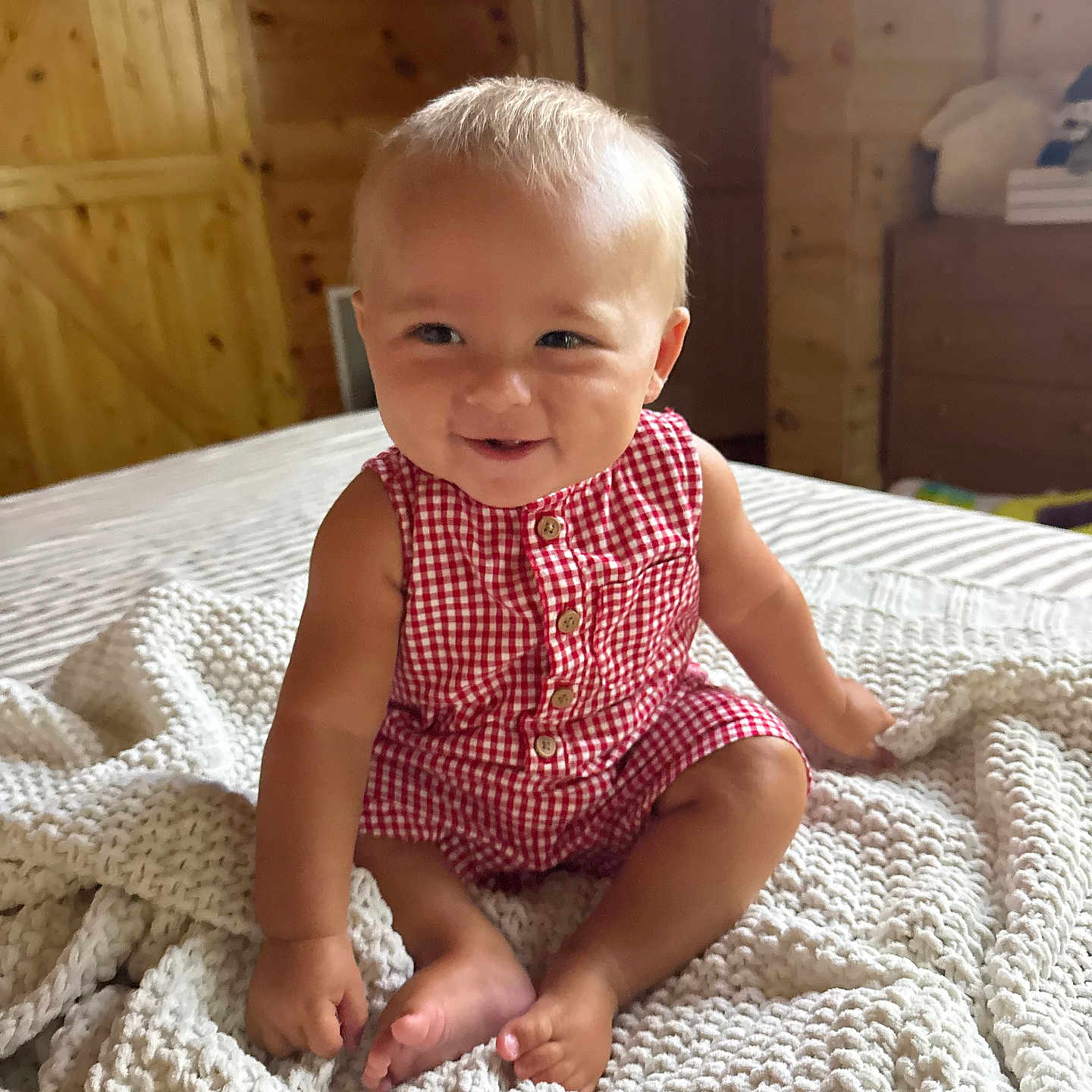 Kohen is registered to the contest to win money with this photo: baby, child, smiling, sitting, red_clothing, romper, blanket, knitted_blanket, wood_wall, indoor, portrait, happy, cute, infant, cozy, furniture, room, natural_light, skin, face