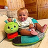 baby, child, clothing, colorful, cozy, cushion, cute, face, feet, happy, indoor, infant, person, pillow, plush, sitting, smiling, tongue_out, toy, wooden_wall