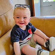 Axel is registered to the contest to win money with this photo: baby, child, smiling, sitting, couch, leather, indoor, shirt, shorts, shoes, socks, sunglasses, bottle, window, door, happy, person, infant, casual_clothing, home