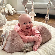 Fiorella is registered to the contest to win money with this photo: baby, infant, smiling, play_mat, plush_toy, soft_toys, pink_clothing, face, hand, floor, sofa, window, wooden_floor, indoor, child, cute, happy, crawling, head, furniture