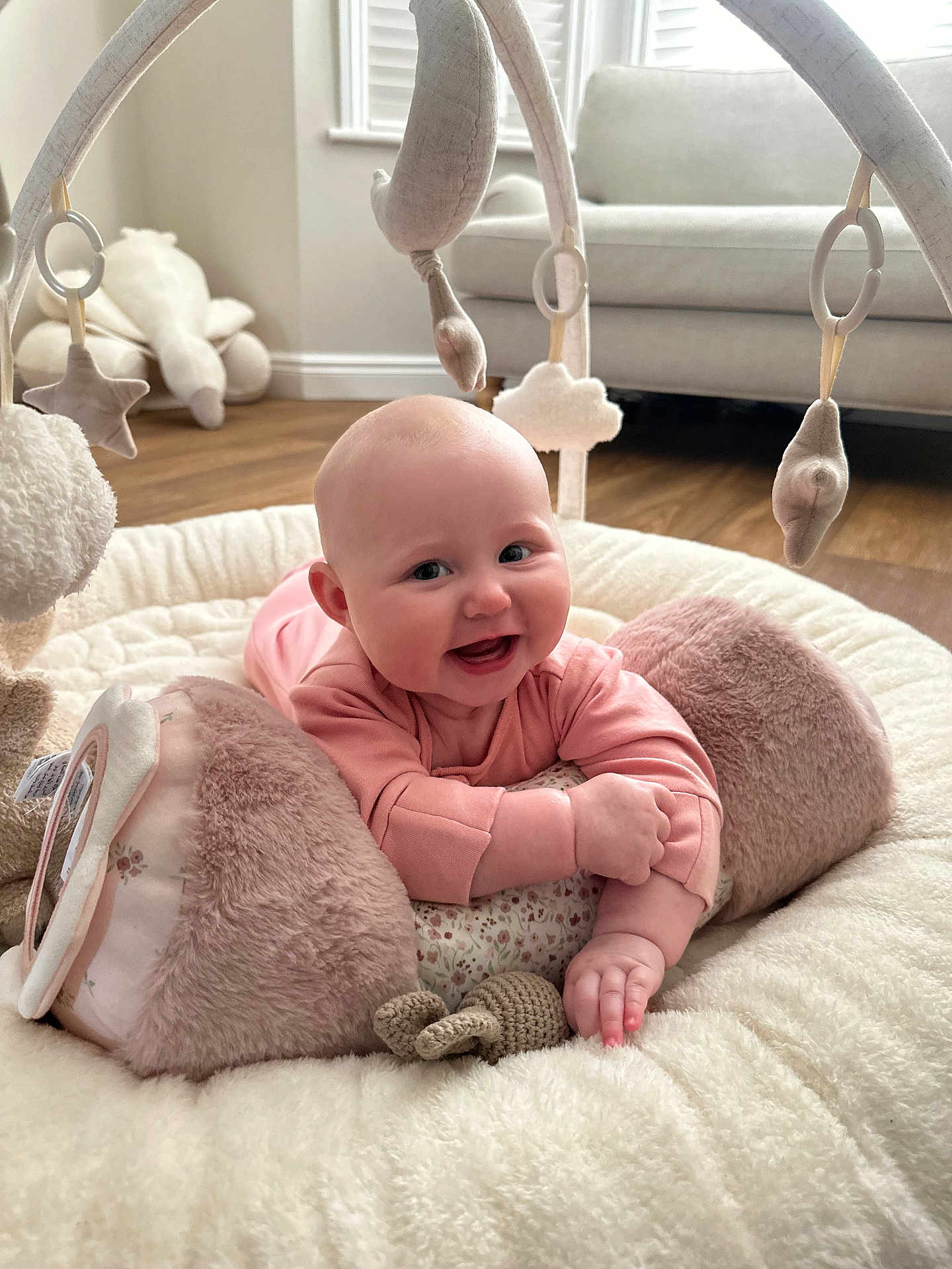 Fiorella is registered to the contest to win money with this photo: baby, infant, smiling, play_mat, plush_toy, soft_toys, pink_clothing, face, hand, floor, sofa, window, wooden_floor, indoor, child, cute, happy, crawling, head, furniture