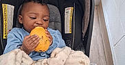Queen is registered to the contest to win money with this photo: baby, child, car_seat, blanket, fruit, yellow_fruit, indoor, window, curly_hair, earring, blue_clothing, soft_texture, floor, wood_floor, person, infant, cute, cozy, natural_light, munching