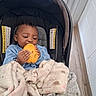 baby, child, car_seat, blanket, fruit, yellow_fruit, indoor, window, curly_hair, earring, blue_clothing, soft_texture, floor, wood_floor, person, infant, cute, cozy, natural_light, munching