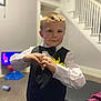 blond_hair, bow_tie, boy, carpet, casual, child, cute, furniture, heart_hands, home, indoor, living_room, person, portrait, smile, stairs, toy, vest, white_shirt, yellow_flower