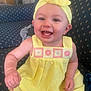 Davina is registered to the contest to win money with this photo: arm, baby, child, clothing, couch, cute, expression, hand, happy, headband, headwear, indoor, infant, patterned_fabric, person, portrait, seated, skin, smile, yellow_dress