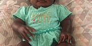 Maame Yaa joined the competition — help win amazing prizes! baby, infant, child, person, clothing, turquoise_outfit, text_on_clothing, bed, bedding, patterned_bedspread, feet, toes, face, hands, cute, sitting, indoor, soft_lighting, portrait, young