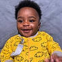 baby, child, smile, yellow_onesie, cloud_pattern, happy, infant, portrait, cute, face, expression, clothing, indoor, person, young_child, hands, black_hair, skin, eyes, soft_background
